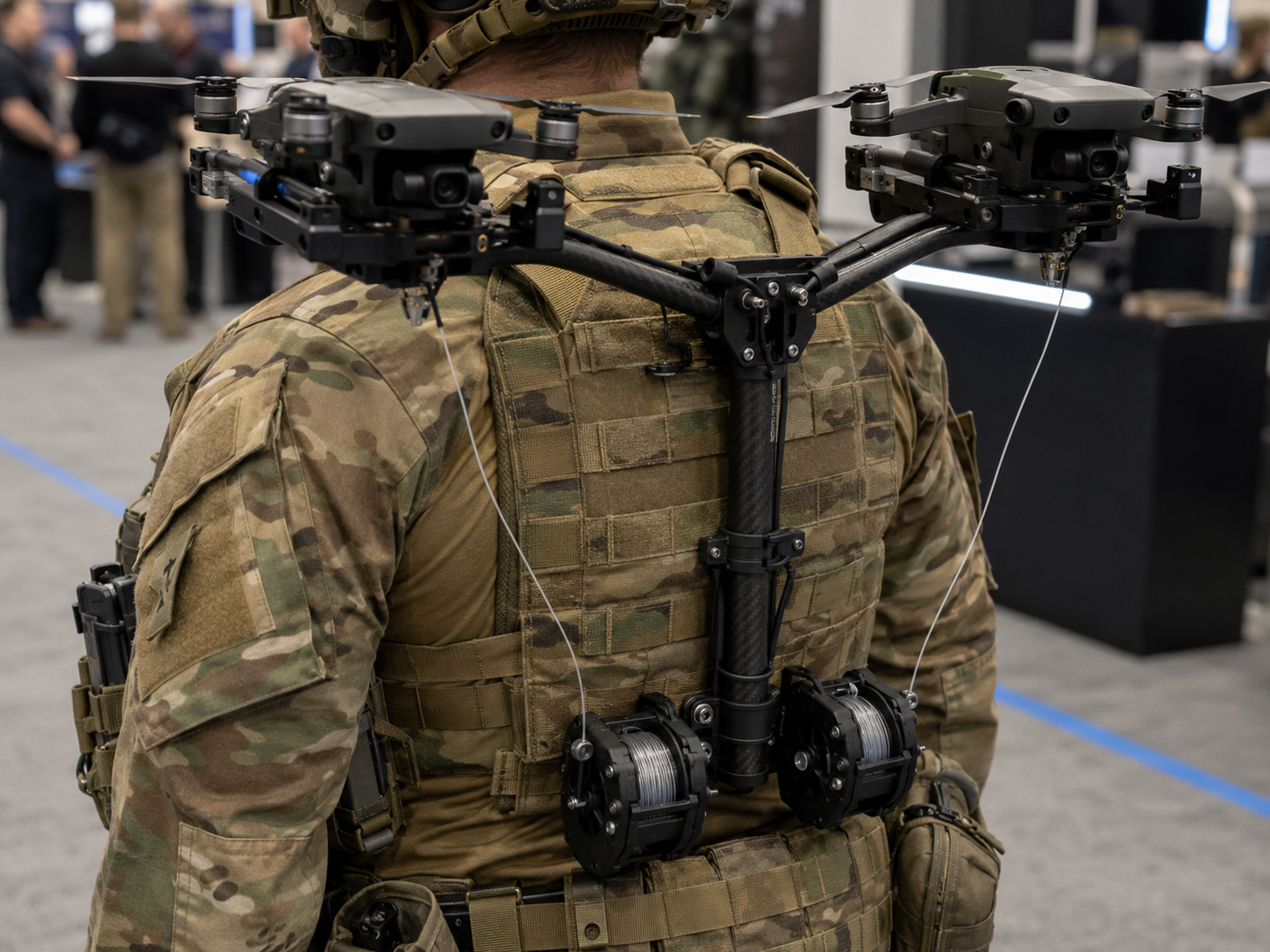 Close view from behind of a soldier's back frame: a carbon mast splits into two arms holding two perched quadcopter drones above the shoulders, with quick-release clamps at each perch and two wound fiber-optic spools mounted at the waist, thin fiber lines running up the mast to each drone.